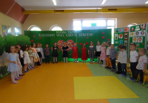 Dzieci stoją na tle dekoracji z serduszek, śpiewają piosenkę.